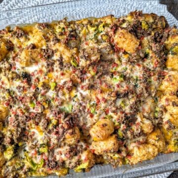 A close-up view shows a freshly baked cracked tater tot hamburger hash casserole in a glass dish. The melted cheese and crispy potato topping create a delicious golden-brown texture for this easy dinner.