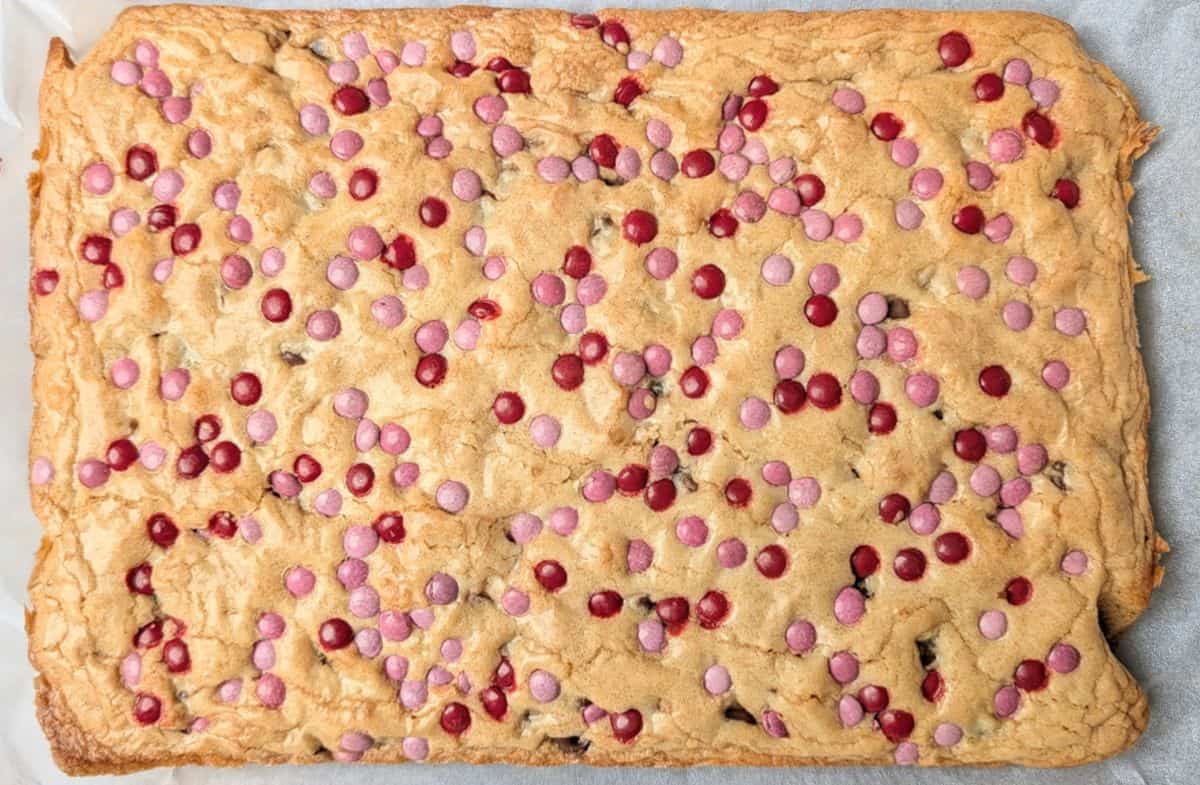 A full baking pan of golden brown Valentine cookie bars topped with melted chocolate candies and sprinkles, fresh out of the oven.