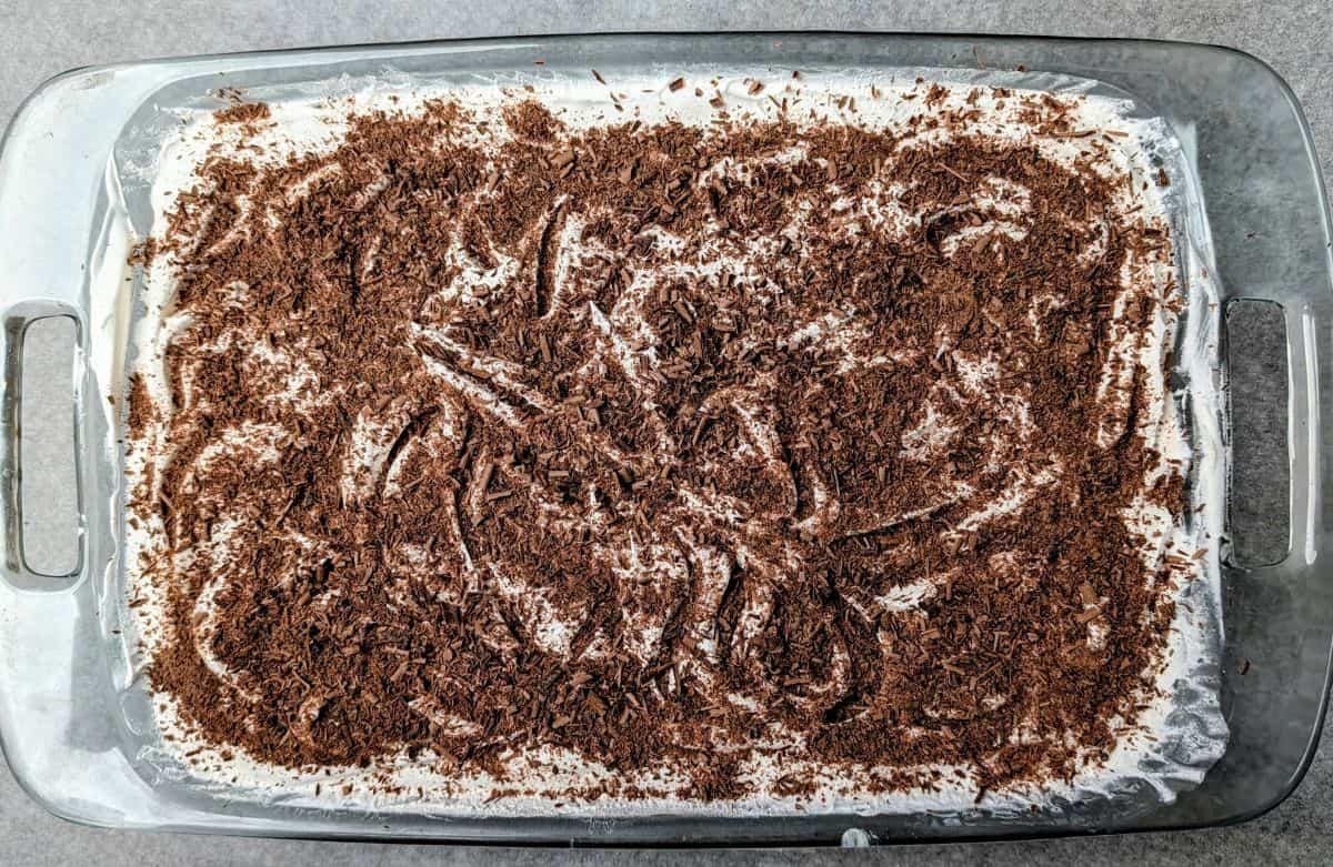 Spreading a thick layer of chocolate shavings over the top of chocolate lasagna in a glass baking dish.