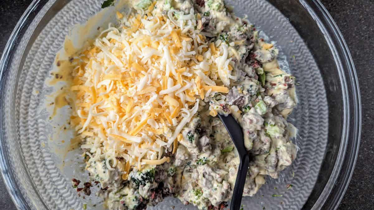 A large mixing bowl is filled with shredded cheese being mixed into the cracked tater tot casserole base. This step ensures every bite of the hamburger hash is packed with gooey, melted goodness.