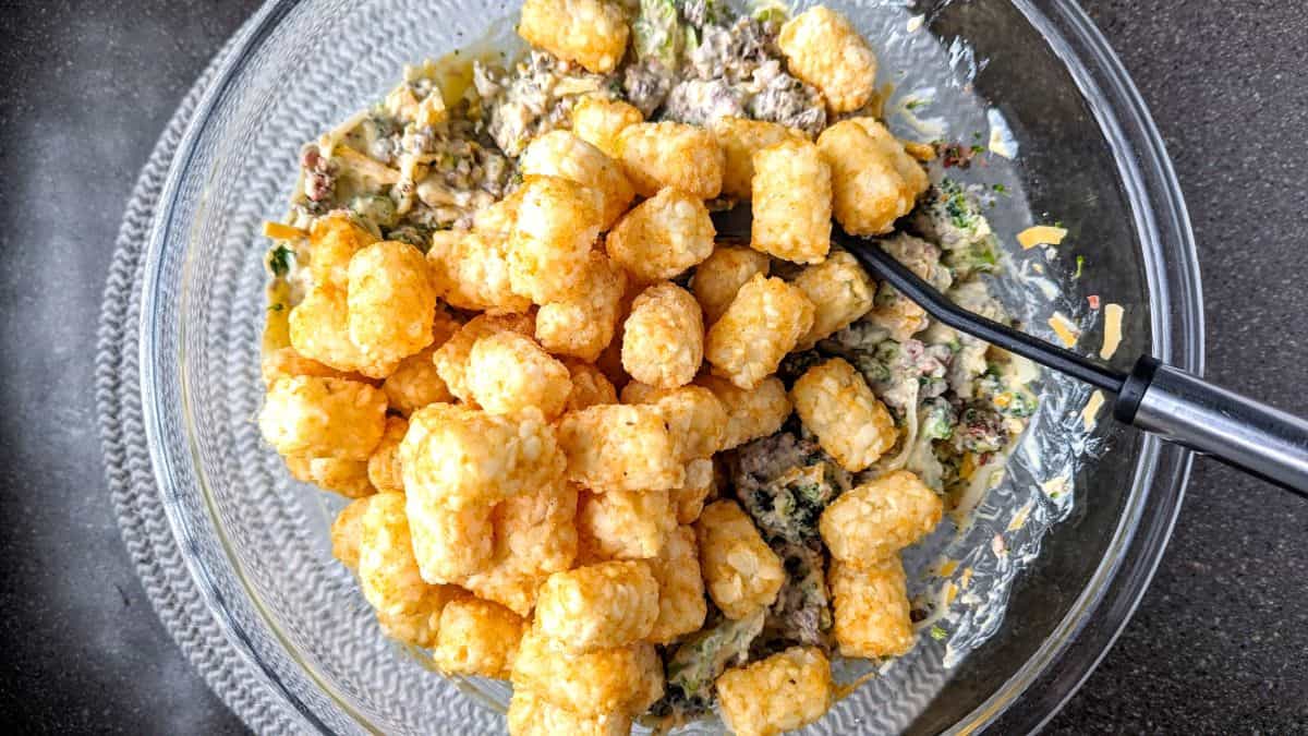 A large spoon mixes golden-brown frozen tater tots into the cheesy beef and broccoli base. Folding in the tots at the very end ensures your cracked tater tot casserole stays perfectly chunky and delicious.