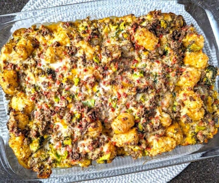A close-up view shows a freshly baked cracked tater tot hamburger hash casserole in a glass dish. The melted cheese and crispy potato topping create a delicious golden-brown texture.