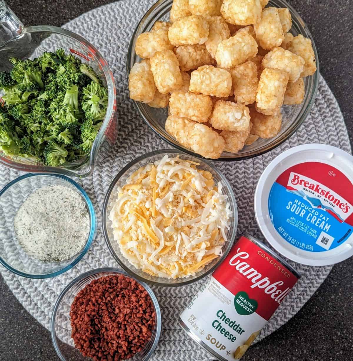 The essential ingredients for a cracked tater tot hamburger hash casserole are laid out in small glass bowls. Fresh broccoli, bacon bits, and shredded cheese surround a pile of frozen tater tots and a can of cheddar soup.
