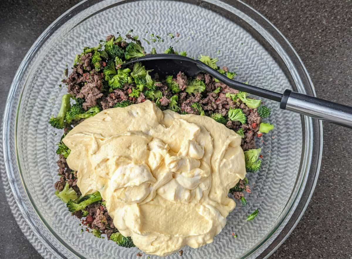 A large mixing bowl contains the creamy beef and broccoli base for a cracked tater tot casserole. The smooth cheddar cheese soup and sour cream mixture is layered over the savory ingredients before mixing.