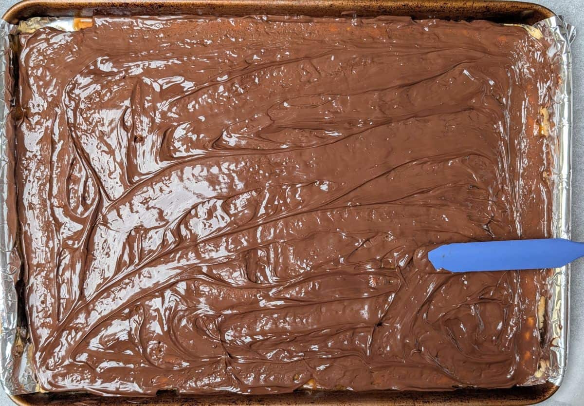 Using a blue spatula to spread a smooth layer of melted chocolate over toffee-coated saltine crackers.