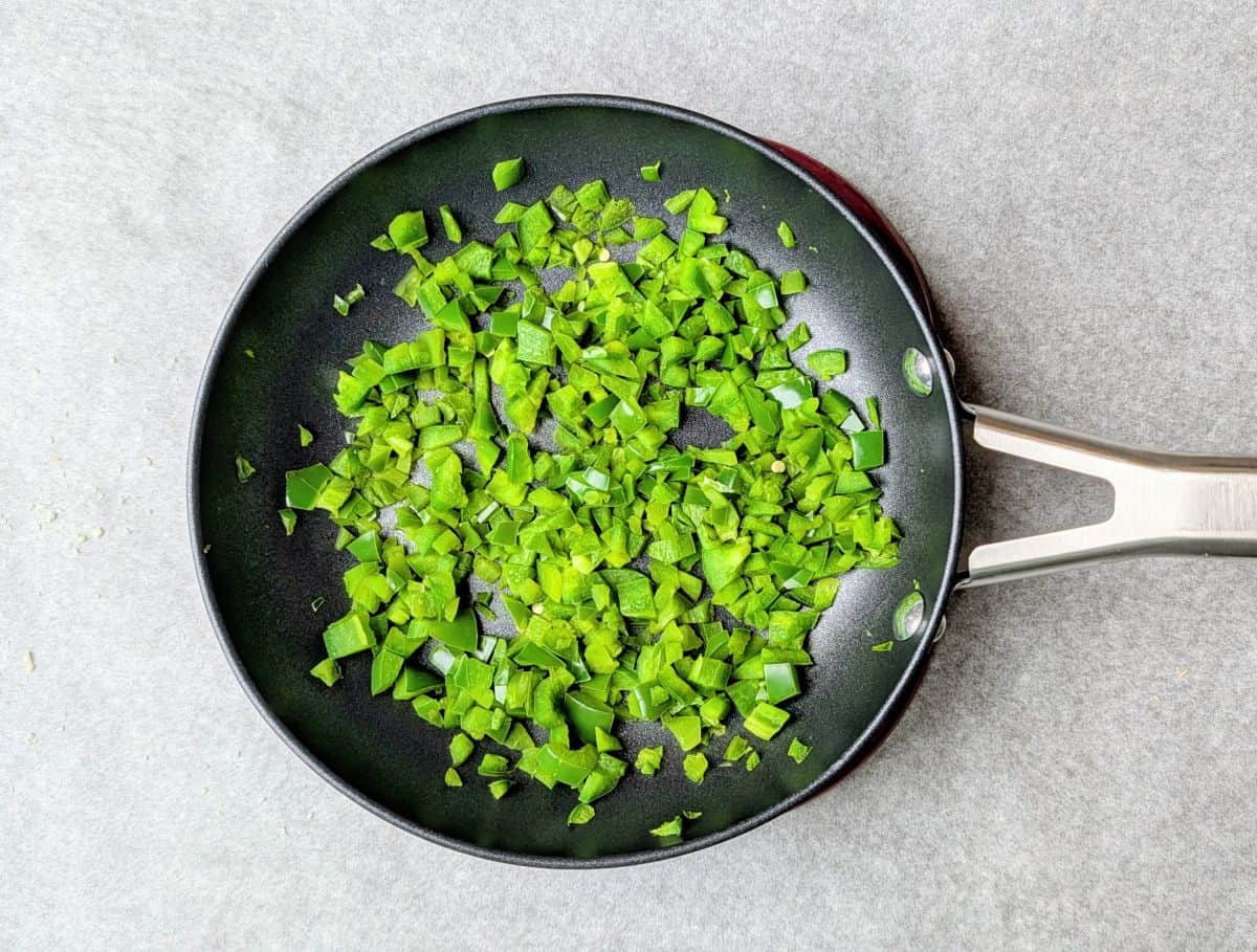 Diced green jalapeños being sautéed in a black frying pan for a homemade jalapeño popper dip recipe.