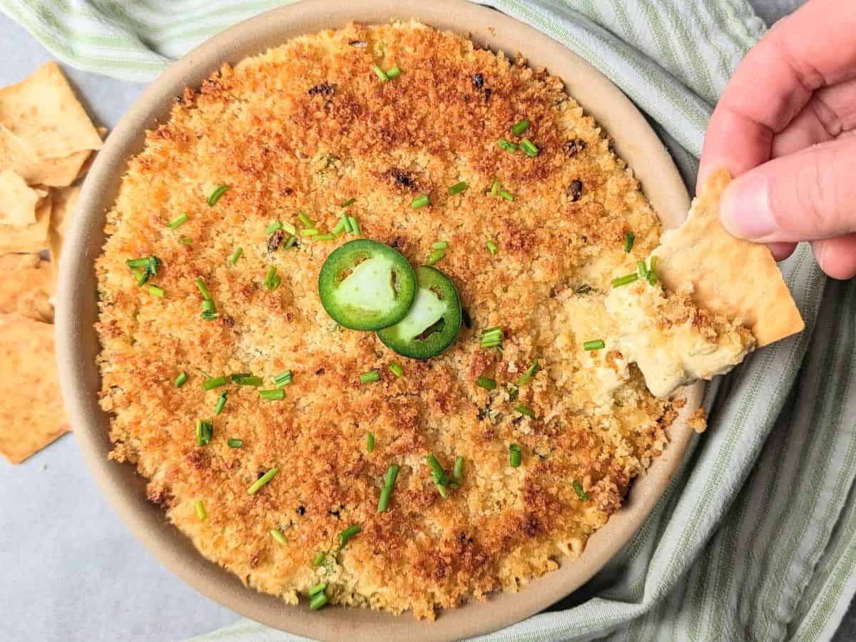 A hand dipping a crunchy pita chip into a warm, golden-brown baked jalapeño popper dip topped with fresh jalapeño slices in a white ceramic dish.