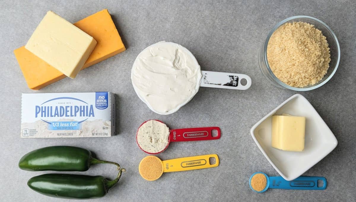 Overhead view of ingredients for jalapeño popper dip including cream cheese, shredded cheddar, fresh jalapeños, and breadcrumbs arranged on a grey surface.