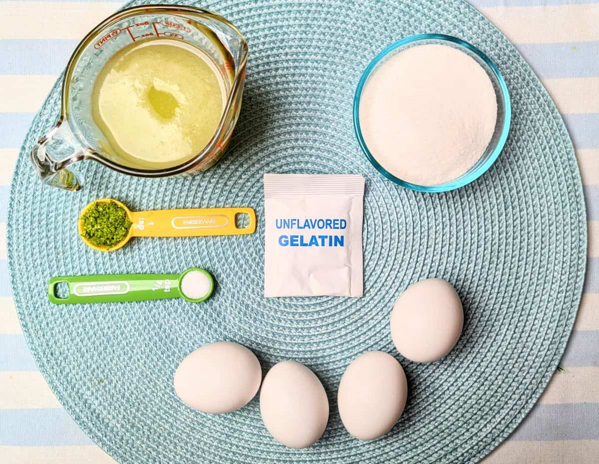 An overhead view of Margarita Pie ingredients on a blue surface, including fresh limes, a citrus juicer, a measuring spoon with unflavored gelatin, a bowl of sugar, and four eggs.
