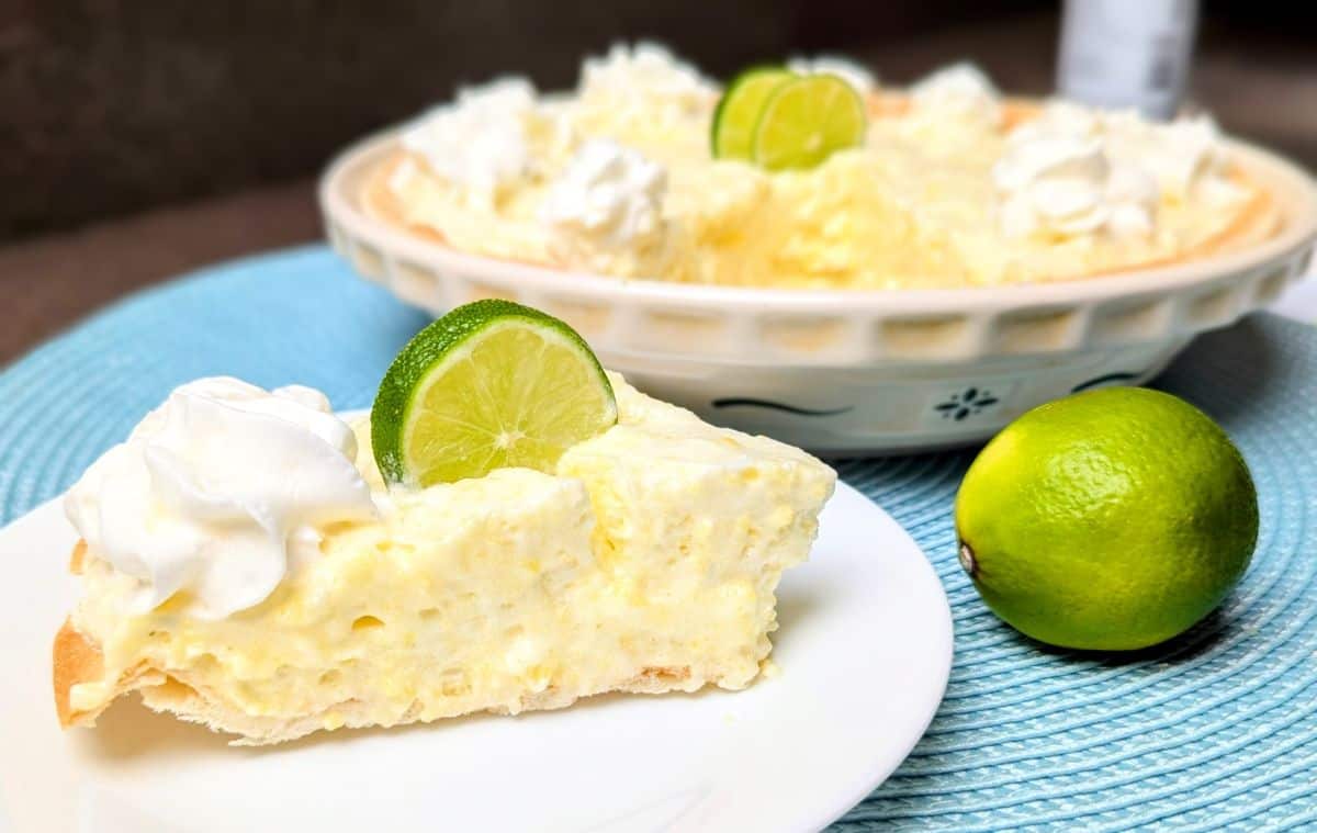 A close-up slice of creamy Margarita Pie on a white plate, showing the fluffy texture and golden crust, garnished with a fresh lime wedge and whipped cream. Two additional slices are visible in the background.