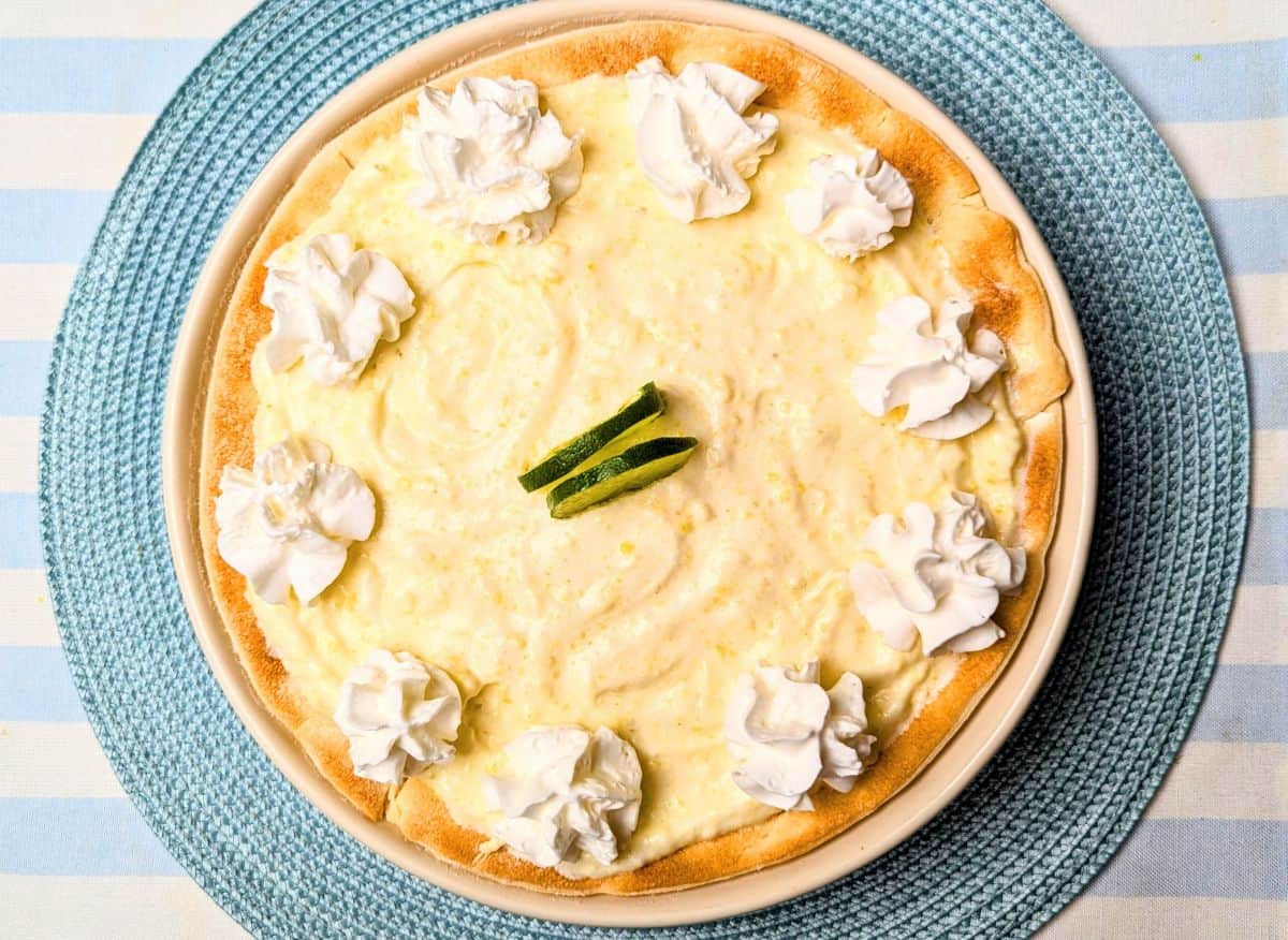 A top-down view of a freshly made Margarita Pie in a golden-brown crust, garnished with a swirl of whipped cream and thin lime slices in the center. The pie is resting on a rustic wooden surface.