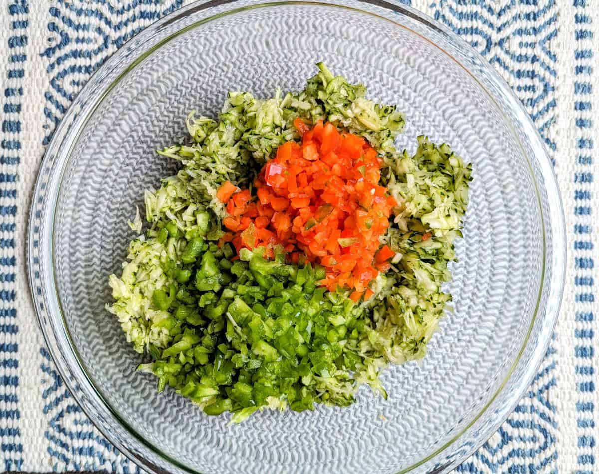 A top-down view of a glass bowl filled with shredded zucchini slaw, mixed with finely diced red and green bell peppers