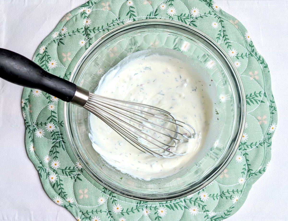 Whisking together sour cream, fresh dill, and spices in a glass bowl to make the dressing for a creamy cucumber salad.