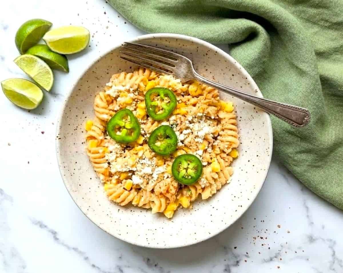 Close-up overhead view of a white bowl filled with Mexican street corn pasta salad, garnished with sliced jalapeños and crumbled cotija cheese on a marble surface.