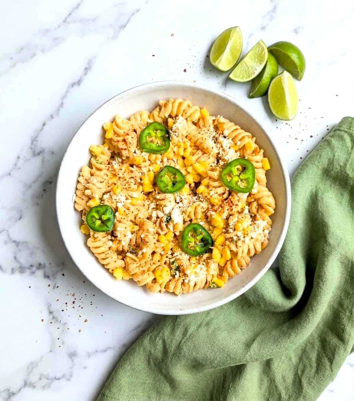 Close-up overhead view of a white bowl filled with Mexican street corn pasta salad, garnished with sliced jalapeños and crumbled cotija cheese on a marble surface.