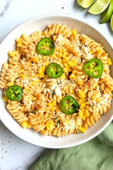 Close-up overhead view of a white bowl filled with Mexican street corn pasta salad, garnished with sliced jalapeños and crumbled cotija cheese on a marble surface.
