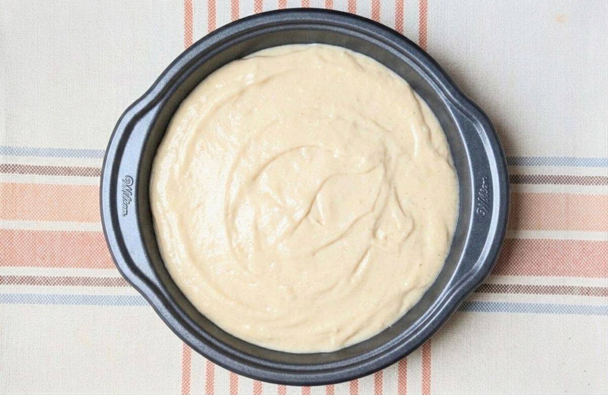 Thick cake batter being smoothed over a layer of fresh sliced peaches in a round baking pan.