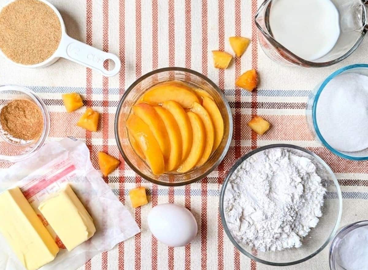 Ingredients for peach upside-down cake, including sliced peaches in a glass bowl, flour, sugar, butter, and spices arranged on a countertop.