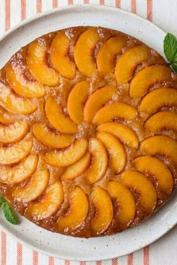 A golden-brown peach upside-down cake on a white plate, featuring a decorative circular pattern of caramelized peach slices on top.