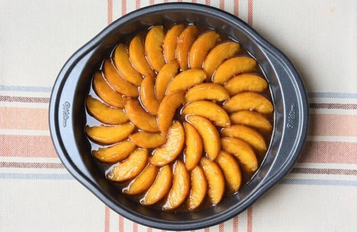 Fresh peach slices arranged in a decorative circular pattern over spiced caramel syrup in a round baking pan.