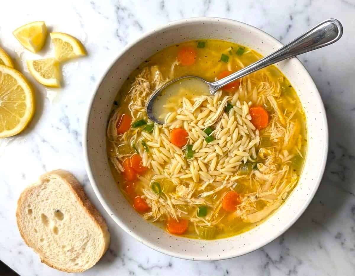 A white bowl of lemon chicken orzo soup with a silver spoon, served with a side of crusty bread and fresh lemon slices.