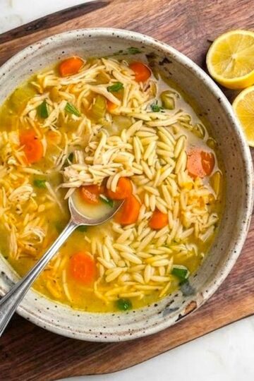 A top-down view of a bowl of lemon chicken orzo soup with fresh herbs and lemon slices, served on a wooden cutting board next to a crusty piece of bread.