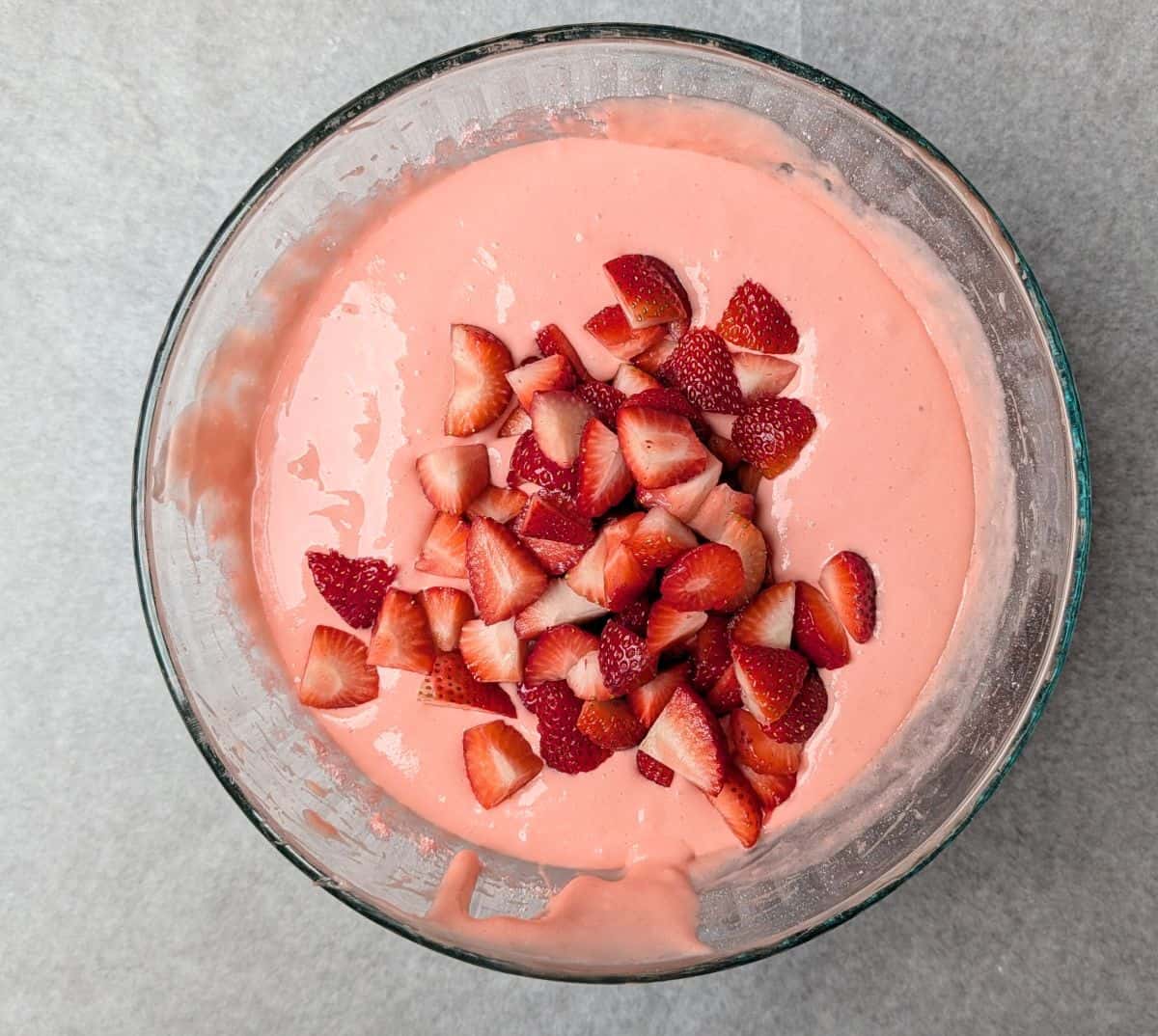 A glass bowl filled with pink strawberry cake batter topped with fresh sliced strawberries, ready to be mixed.
