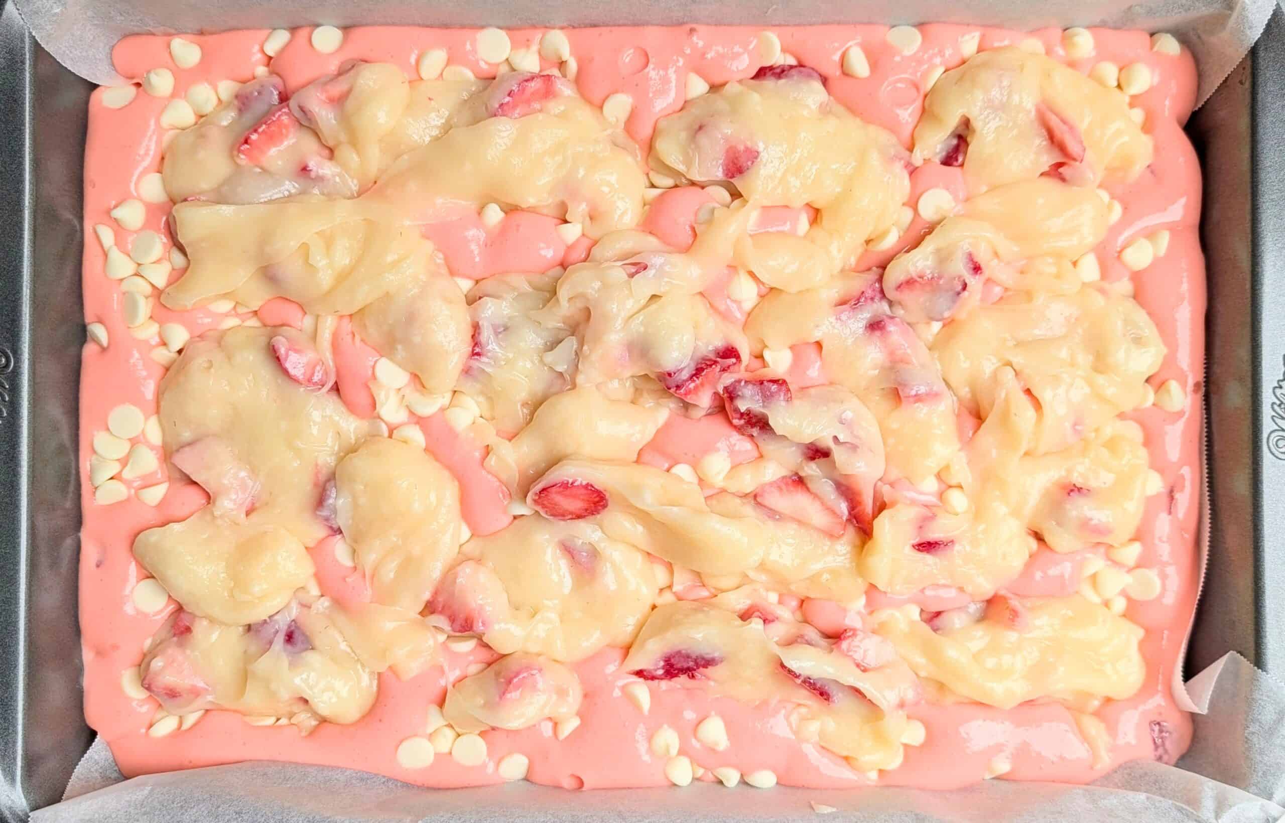 A top-down view of pink strawberry earthquake cake batter in a rectangular pan, topped with white chocolate chips and swirls of cream cheese filling.