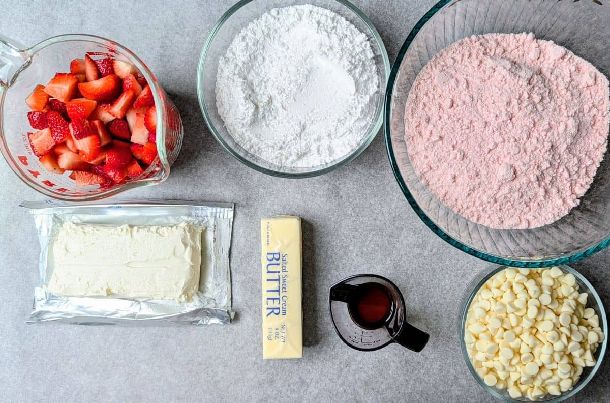 An overhead view of the ingredients for strawberry earthquake cake, including pink cake mix, cream cheese, butter, powdered sugar, white chocolate chips, and fresh strawberries.