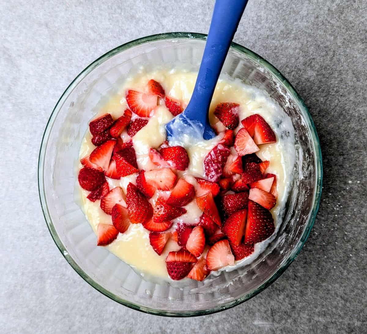Stirring fresh sliced strawberries into a white cream cheese filling in a glass bowl with a blue spatula.