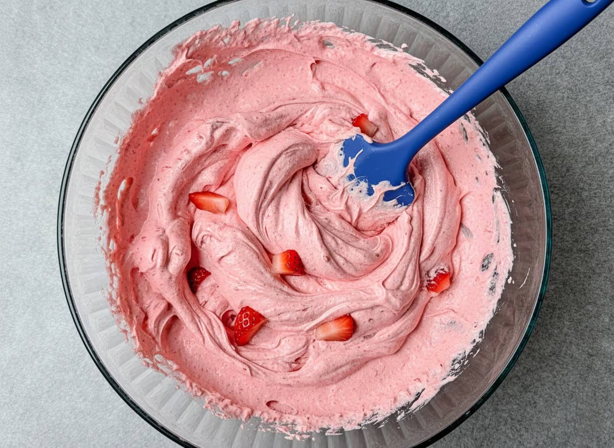 Using a blue spatula to fold fresh chopped strawberries into the creamy pink base of a strawberry fluff salad.