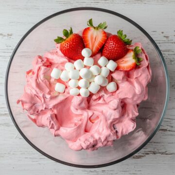 A glass serving bowl filled with creamy pink strawberry fluff salad, topped with three fresh strawberry halves for garnish.