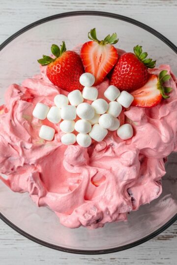 A glass serving bowl filled with creamy pink strawberry fluff salad, topped with three fresh strawberry halves for garnish.