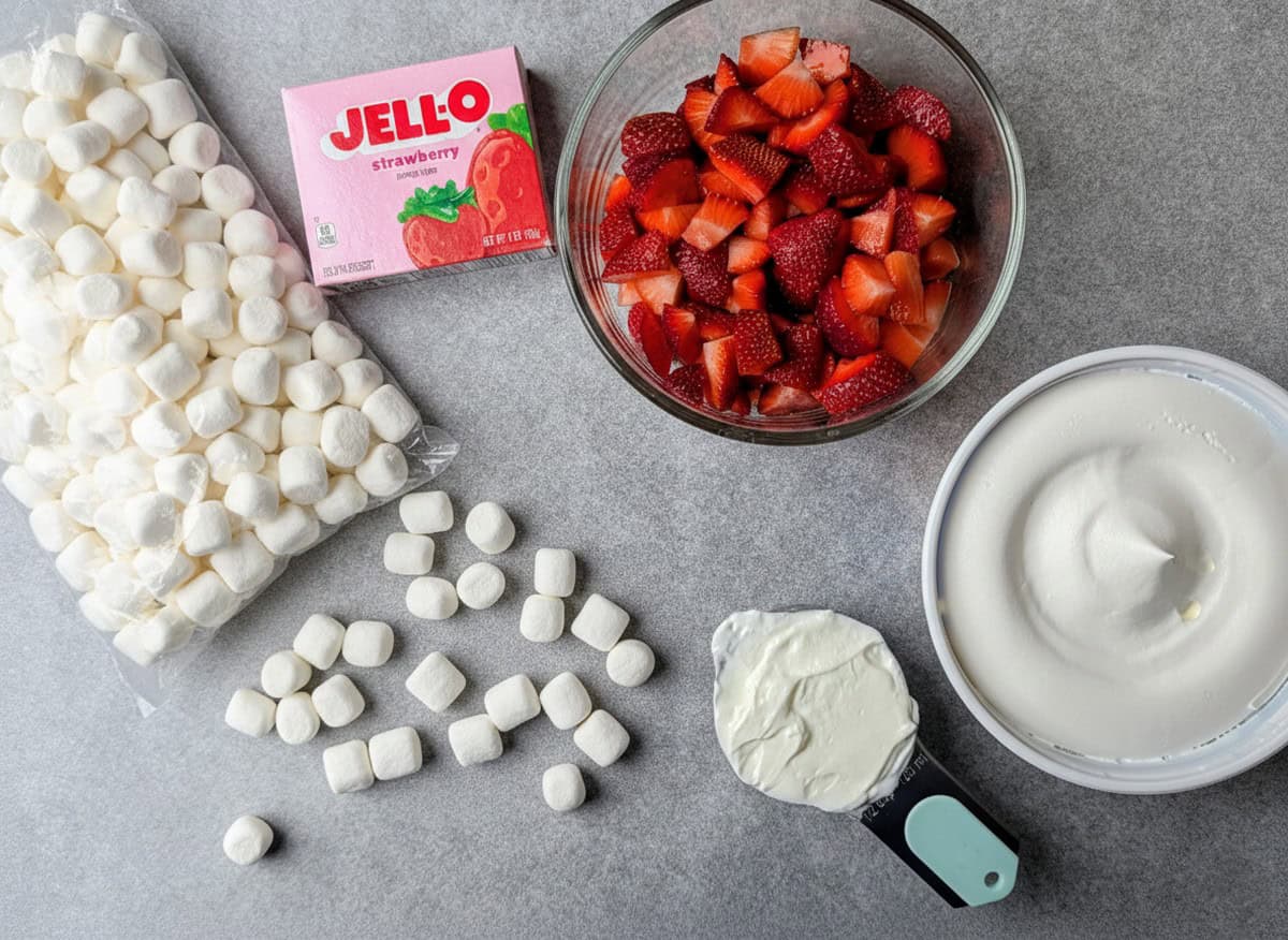 Ingredients for strawberry fluff salad including a box of strawberry Jello, mini marshmallows, fresh sliced strawberries, and a bowl of whipped topping.