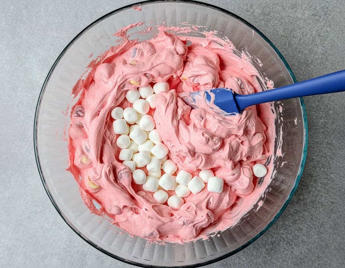 A blue spatula stirring mini marshmallows into the creamy pink base of a strawberry fluff salad in a glass mixing bowl.