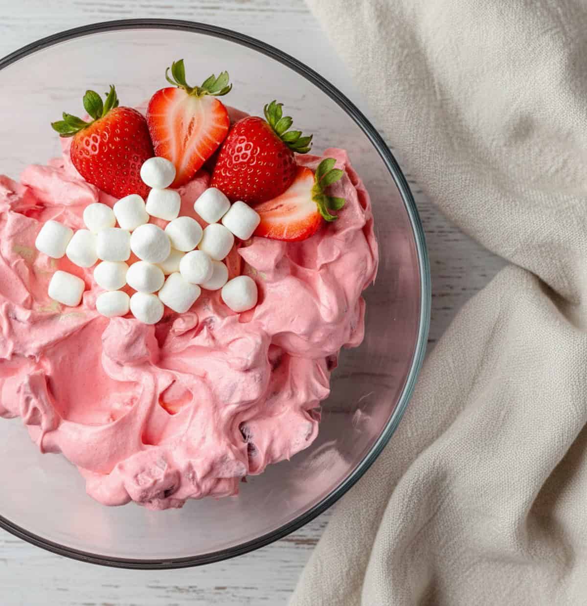 A glass bowl of creamy strawberry fluff salad topped with fresh strawberry halves and mini marshmallows, served on a grey linen napkin.
