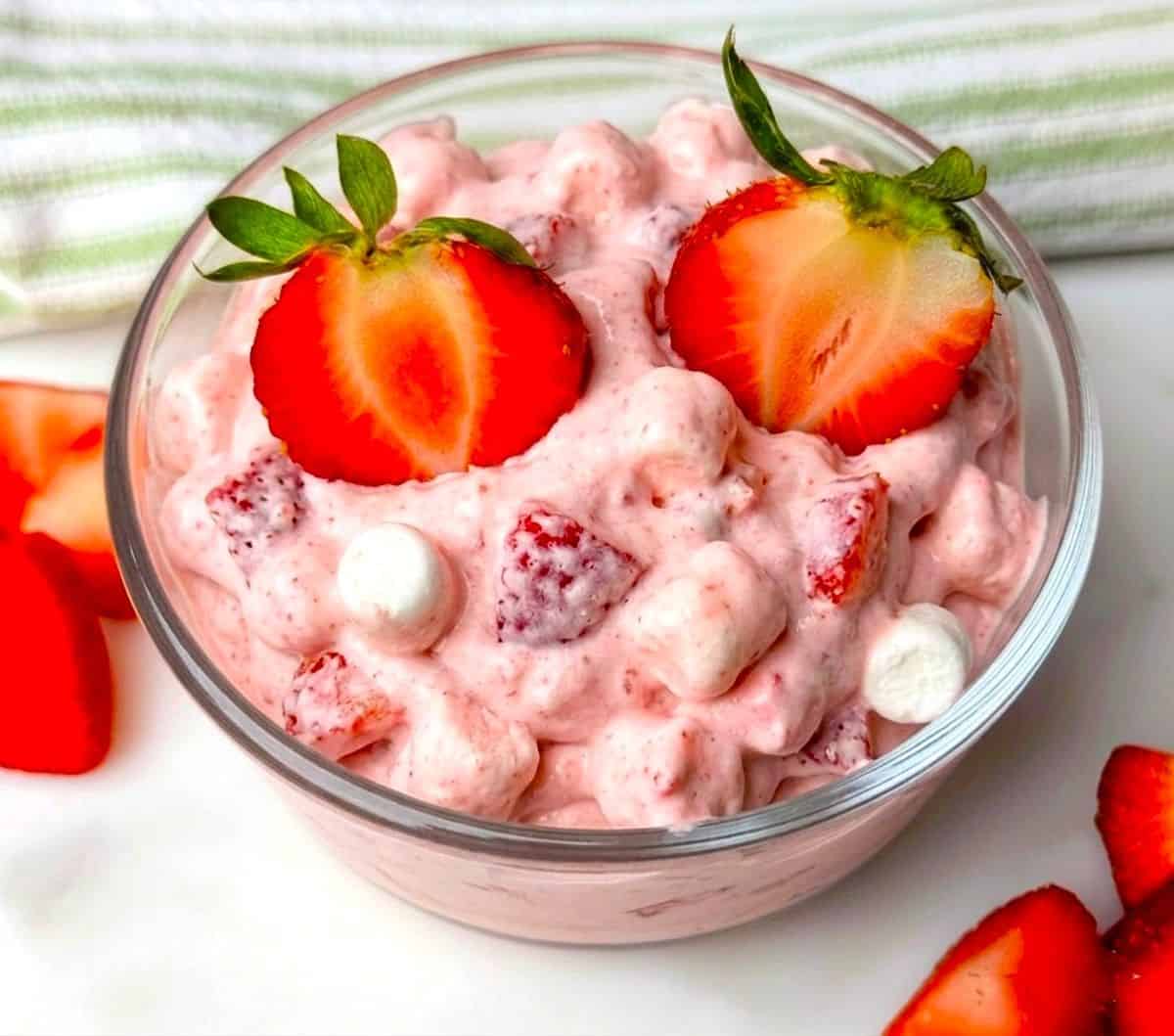 A glass serving bowl filled with creamy pink strawberry fluff salad, topped with three fresh strawberry halves for garnish.