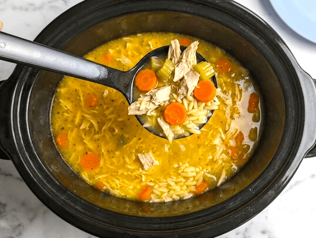 A top-down view of a slow cooker filled with lemon chicken orzo soup, featuring a metal ladle scooping up shredded chicken, carrots, and orzo pasta.
