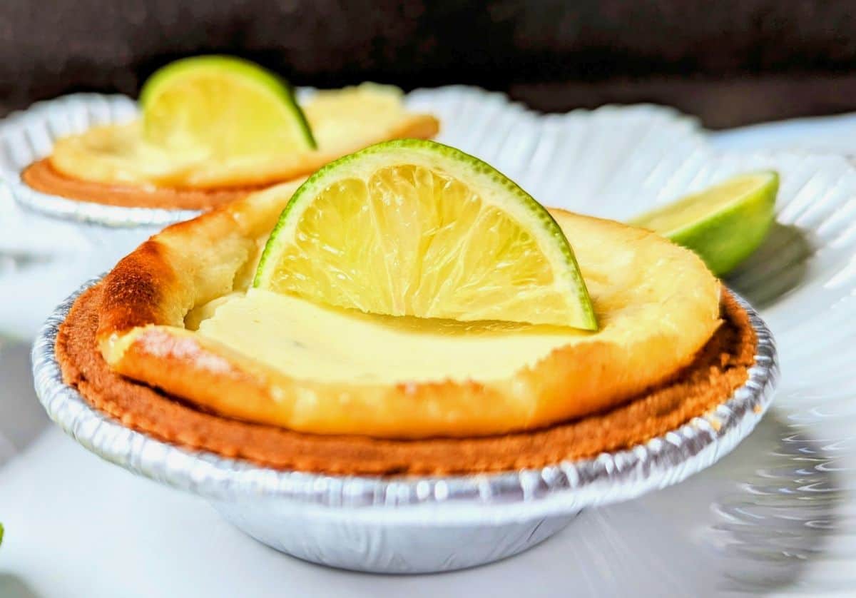 Close-up of a mini key lime cheesecake in a graham cracker crust, garnished with fresh lime slices and zest.