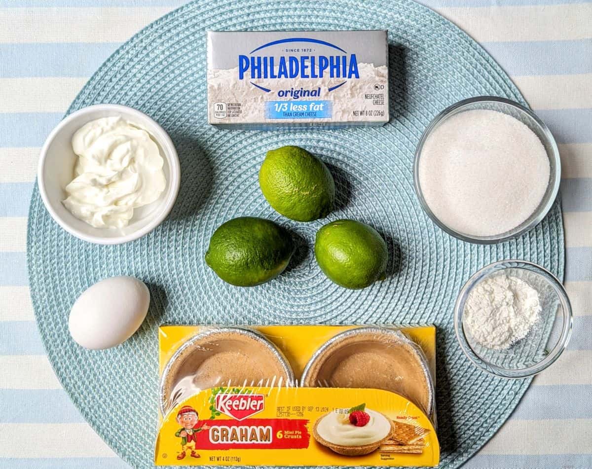 Overhead view of the ingredients needed for mini key lime cheesecakes, including fresh limes, Philadelphia cream cheese, and sugar.