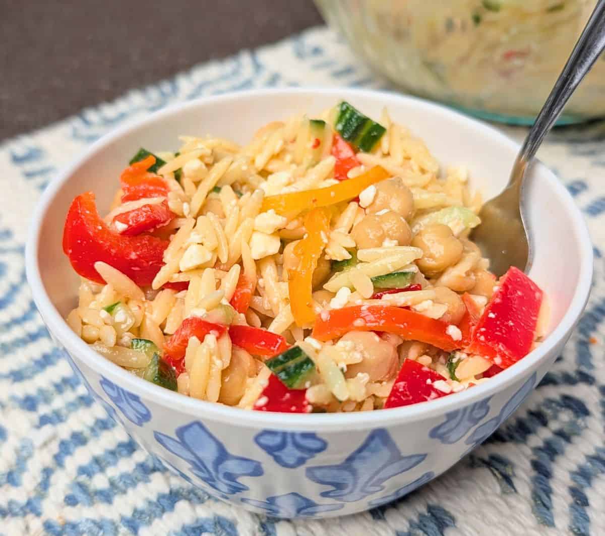 A close-up of Orzo Greek Salad in a white bowl with a fork, featuring orzo pasta, crumbled feta cheese, diced tomatoes, and cucumbers.
