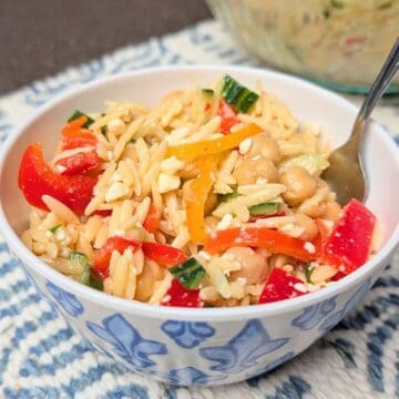 A close-up of Orzo Greek Salad in a white bowl with a fork, featuring orzo pasta, crumbled feta cheese, diced tomatoes, and cucumbers.