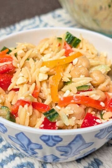 A close-up of Orzo Greek Salad in a white bowl with a fork, featuring orzo pasta, crumbled feta cheese, diced tomatoes, and cucumbers.