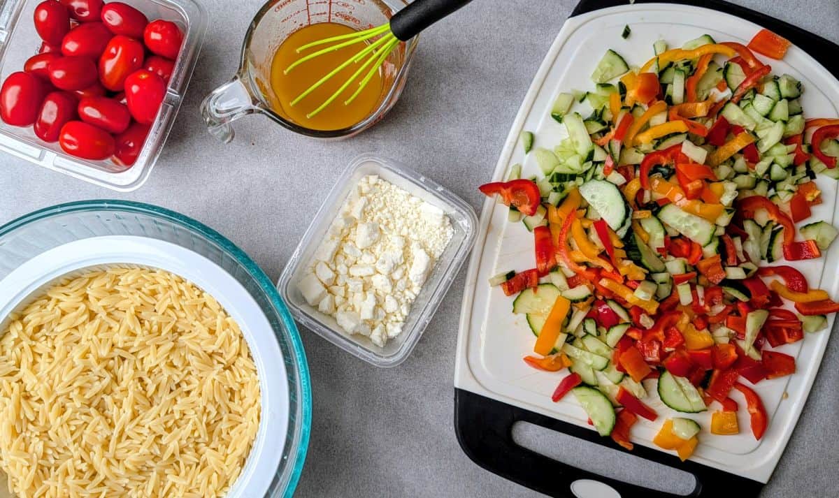 Overhead shot of raw Orzo Greek Salad ingredients on a white marble surface, including a bowl of cooked orzo pasta, fresh diced tomatoes, feta cheese, cucumbers, and a glass measuring cup with homemade Mediterranean dressing and a whisk.