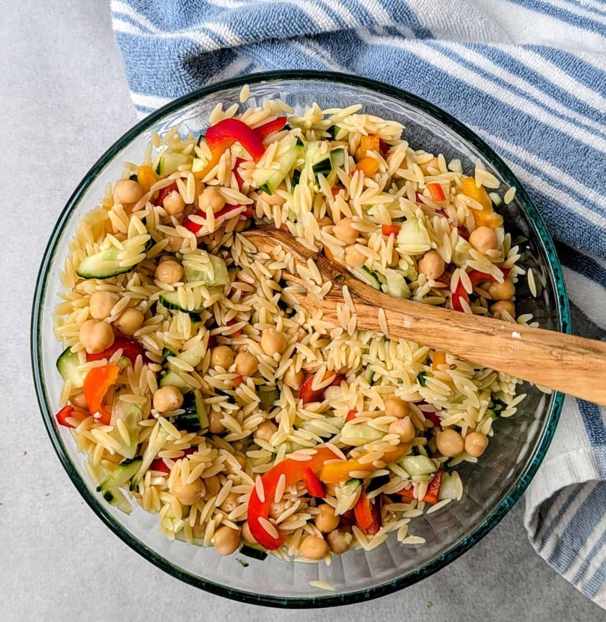 Overhead shot of a freshly mixed Greek orzo pasta salad in a clear glass bowl, featuring cooked orzo, diced tomatoes, cucumbers, and chickpeas, resting on a blue striped linen with a wooden spoon.