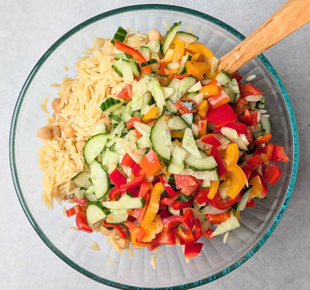 Tossing the colorful base ingredients of a Greek orzo pasta salad, featuring bright red tomatoes and crisp cucumbers, in a clear glass mixing bowl.
