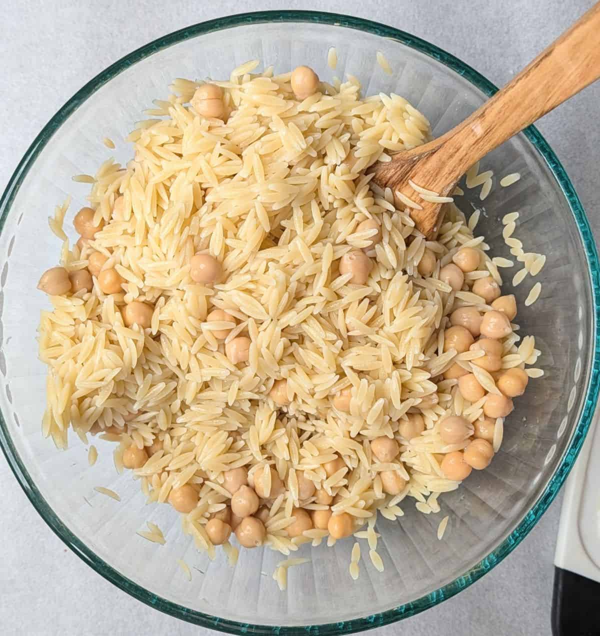 Overhead view of cooked orzo pasta and hearty chickpeas being stirred with a wooden spoon in a glass mixing bowl for an easy Greek orzo salad.