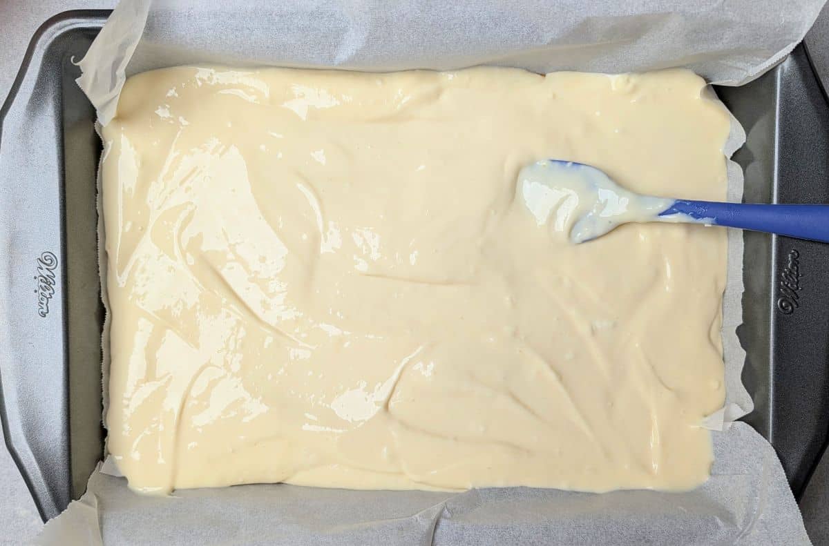 A spatula spreading the smooth filling over a prepared crust for lemon cream cheese bars.