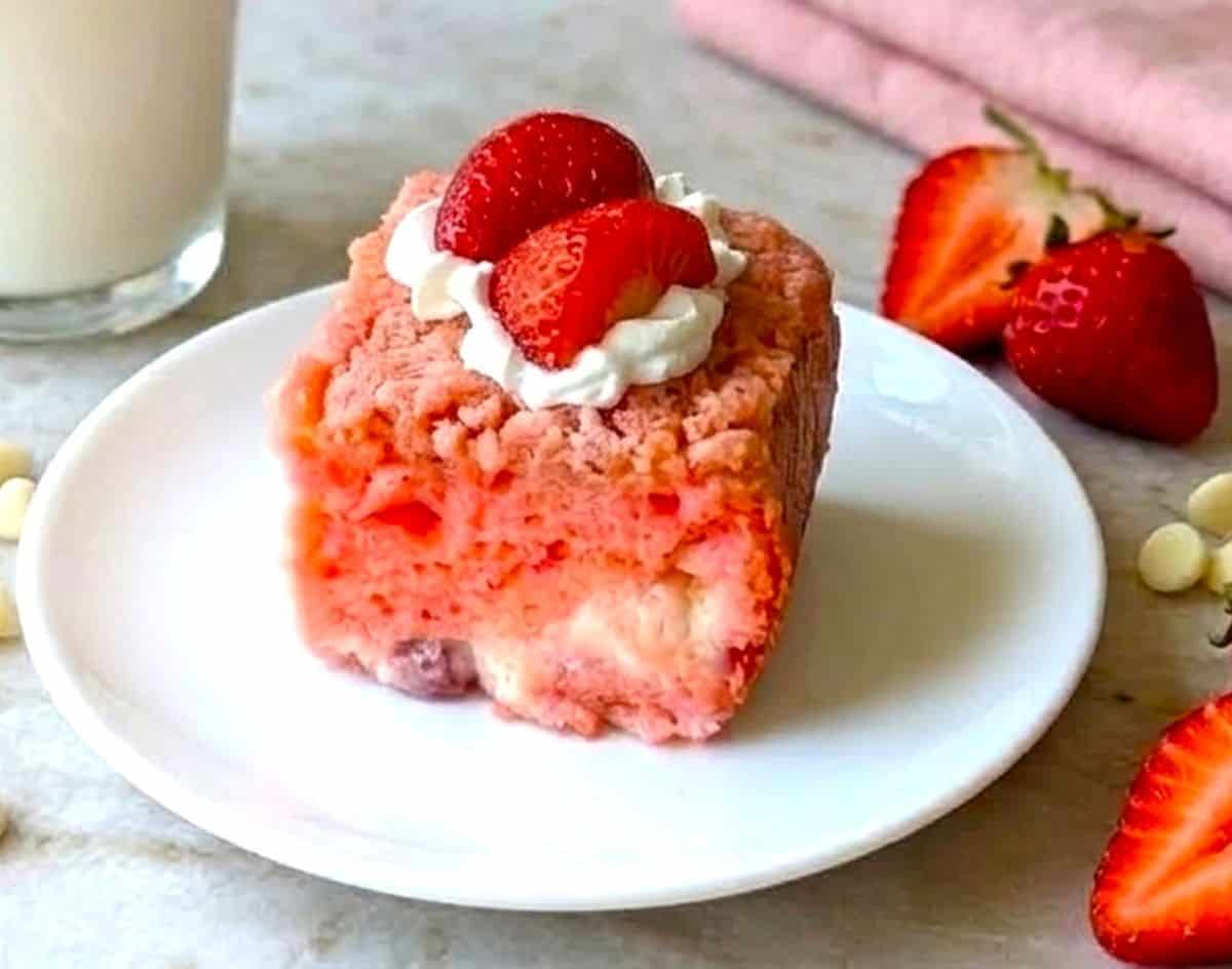 A square slice of pink strawberry earthquake cake on a white plate, topped with whipped cream and a fresh strawberry, served with a glass of milk.