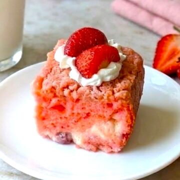 A slice of pink Strawberry Earthquake Cake on a white plate, topped with a dollop of whipped cream and a fresh strawberry half, with a fork nearby.